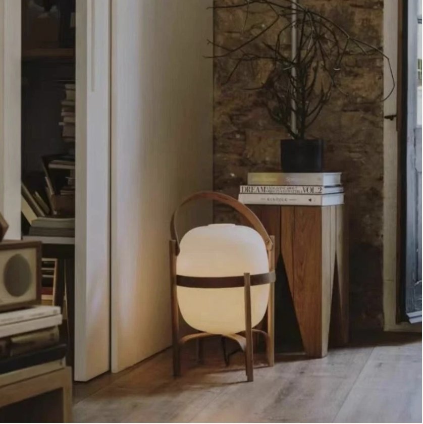 Lampe lanterne baladeuse en verre opalin et bois, poignée en cuir, posée sur un tabouret dans un intérieur cosy